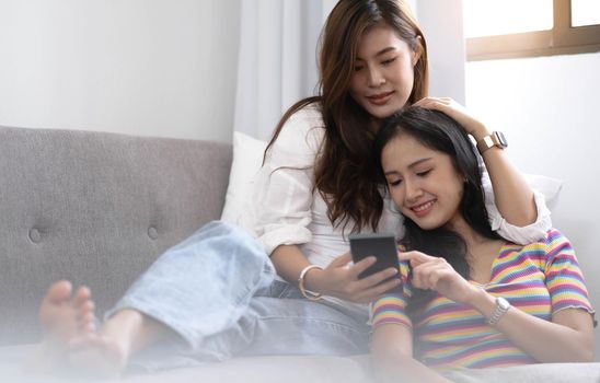 Young beautiful Asian women lesbian couple lover using smartphone video call online in living room on sofa at home with smiling face.Concept of LGBT sexuality with happy lifestyle together.