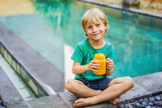 Boy drinking juicy smoothie from mango in glass mason jar with steel straw on the background of the pool. Healthy life concept, copy space