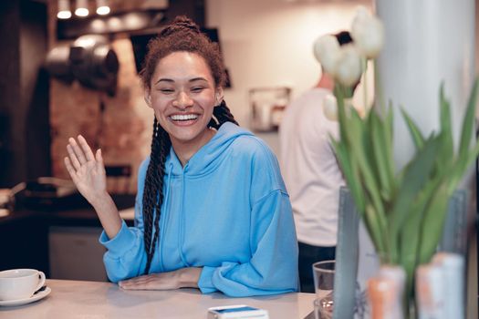 Pretty mixed-race young female bartender with piercing smiling at counter and waving hand.
