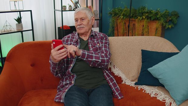 Senior old grandparent works on mobile phone, sends messages, makes online purchases at home sofa
