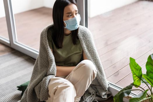 Covid 19 and pandemic concept. Sad ill asian girl in medical face mask self-quarantine at home, looking outside window with upset yearning face expression