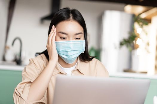 Young sick asian girl in medical face mask using laptop, working from home on quarantine