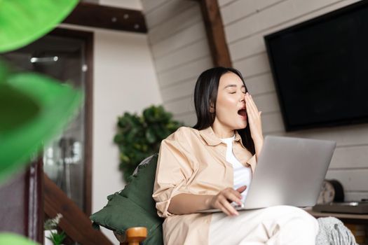 Tired asian girl yawns during work on laptop, sits at home, works remotely from her living room