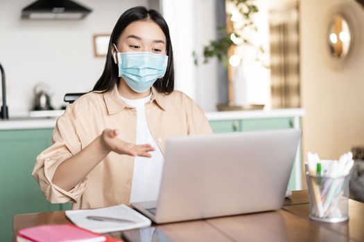 Young asian girl talking at laptop in medical mask, sitting at home, work or study remotely