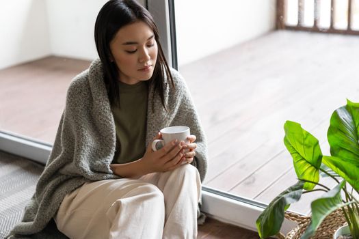 Young sad asian woman sitting on floor near balcony and looking at tea mug thoughtful