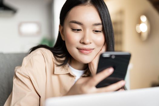 Portrait of asian girl sitting with laptop, checking her phone and smiling, browsing websites on smartphone