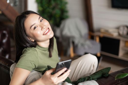 Dreamy smiling asian woman using mobile phone, sitting and relaxing at home