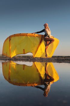 Attractive stylish young caucasian woman in cap sunglasses and kitesurfer outfit standing next to sandy shore next to her kite at sunset reflecting in the water