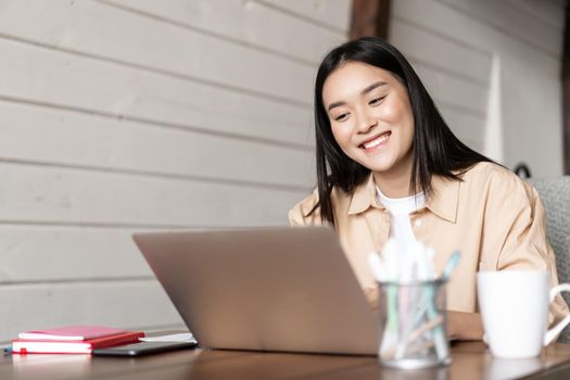 Smiling asian girl looks at laptop screen, works from home, listens webinar or attends online lecture, university classes on remote