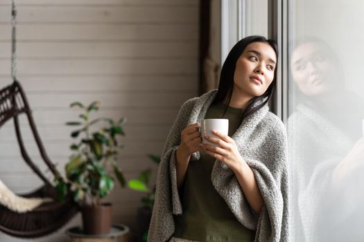 Romantic asian woman wrapped in blanket, leaning on window and looking outside, drinking hot coffee
