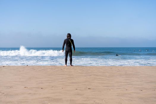 One man dressed black wetsuit standing ocean beach rear view looking on horizon