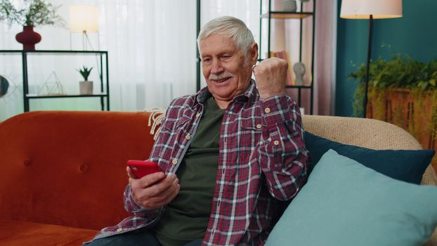 Overjoyed senior grandfather man hold smartphone, excited about mobile app sport bet bid win at home