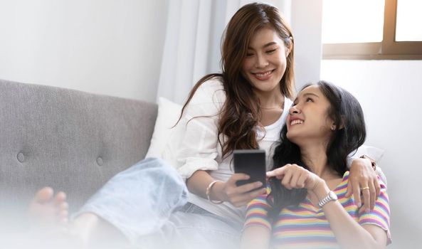 Young beautiful Asian women lesbian couple lover using smartphone video call online in living room on sofa at home with smiling face.Concept of LGBT sexuality with happy lifestyle together.