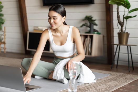 Young asian woman connect to online fitness instructor on laptop, workout via video chat at home, sitting on floor mat