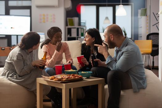 Multiethnic group of coworkers having conversation at office celebration. Man and women talking and having fun with alcoholic drinks, pizza and chips at party after work hours.