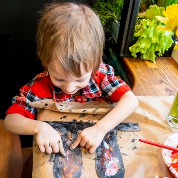 boy draws paper rabbit ears with finger paints. Preparation for Easter: a child paints paper rabbit ears with paints in the kitchen among fresh greenery, flowers and painted eggs