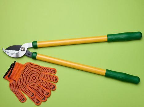 Large garden pruner for pruning branches on trees and orange textile gloves on a green paper background, top view
