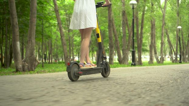 Young woman in a white dress rides a sharing electro scooter in the park. Bottom view.