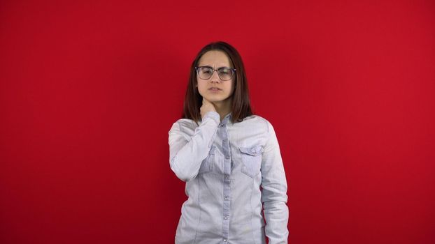 A young woman with glasses has a pain in her neck, she holds her neck with her hand. Shooting on a red background.