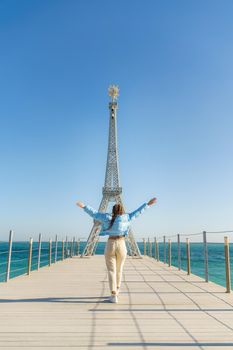 Large model of the Eiffel Tower on the beach. A woman walks along the pier towards the tower, wearing a blue jacket and white jeans.