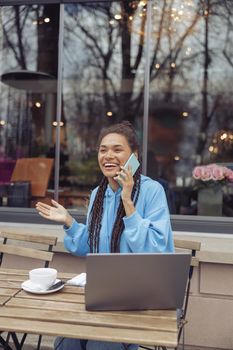 Pretty smiled young mixed-race female talking on cellphone while having coffee outdoor at laptop.