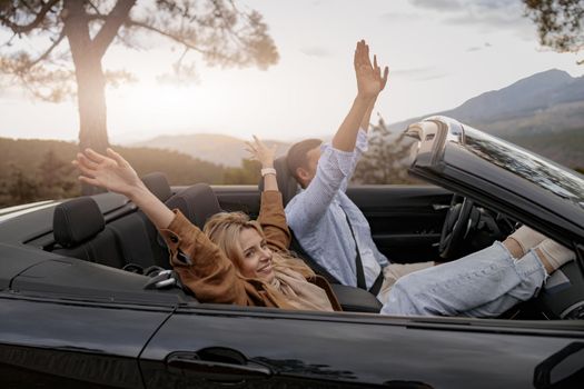 Side view of cheerful boyfriend and girlfriend sitting in convertible car in mountains