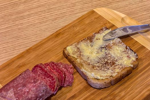 Freshly spread a slice of bread with butter, sliced salami and knife on a cutting board