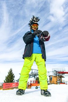 Attractive youg man tourist in funny mohawk hat, green pants, ski glasses mask and black mittens on a background of blue sky. Winter holiday and pastime concept.