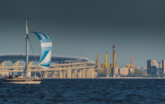Russia, St. Petersburg, 20 May 2022: Sailing boats in the background of the new highway in sunset light, cranes in the port in the background