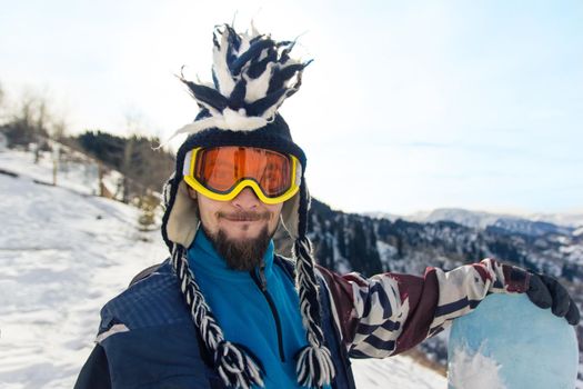 Funny bearded man snowboarder portrait in mohawk hat enjoys the ski resort in the mountains