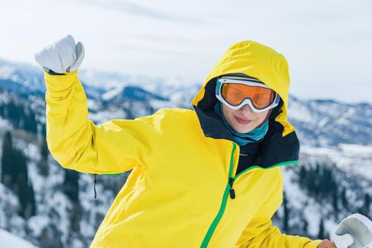 Beautiful youg woman tourist in yellow jacket, ski glasses mask and white mittens on a background of mountains looking at the camera. Winter holiday and pastime concept.