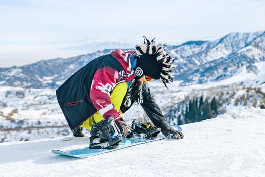 Young man snowboarder in fanny hat sits in the snow and fastens fastenings on a snowboard before the descent