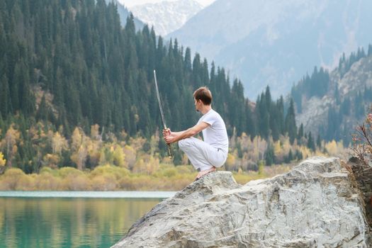 A wise man sits on a stone and holds a sword in his hands. Mountain Lake background.