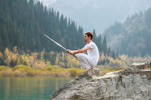 A wise man sits on a stone and holds a sword in his hands. Mountain Lake background