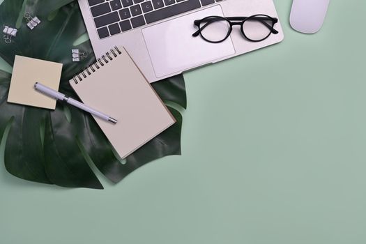 Laptop computer, notepad, glasses and monstera leaf on green background. Top view.