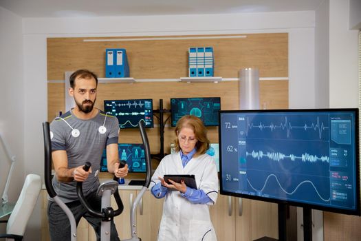 Male athlete who makes a physical effort on the stepper and has electrodes attached to him and the doctor who follows the evolution of his tablet