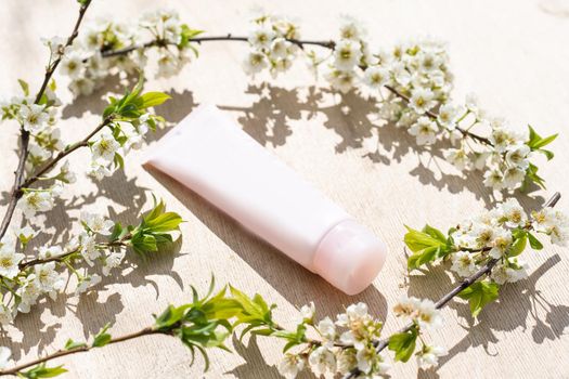 Blank white cosmetics tube and spring flowering tree branch with white flowers on pastel background. Front view