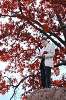 A man with a book in his hands stands against the background of a tree with red leaves
