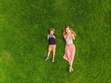 Mom and son are lying on the grass in the park. Mom looks at the phone, son looks at the tablet. Photos from the drone, quadracopter