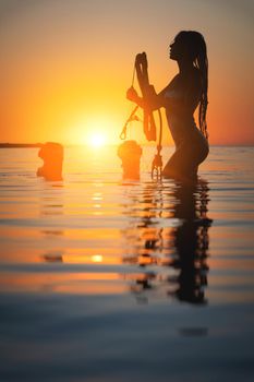 Silhouette frame. A young slim sexy female kitesurfer with a plank and a kiteboard stands in the water in the shallow water at sunset. Water sports. Stylized frame