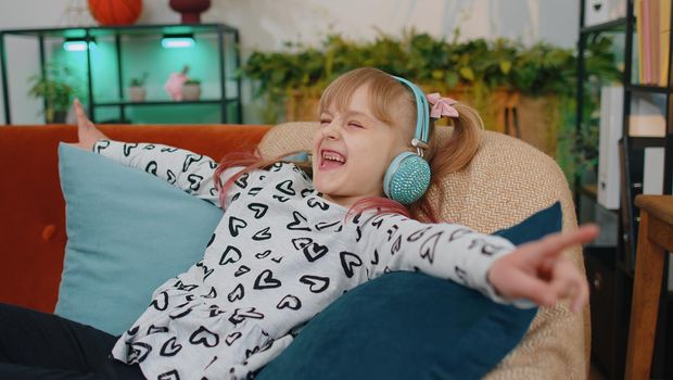 Little children girl lying on sofa, wearing wireless headphones, listening to music, singing song