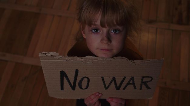 Afraid homeless toddler girl sitting holding inscription No War, hiding from bombing attack at home