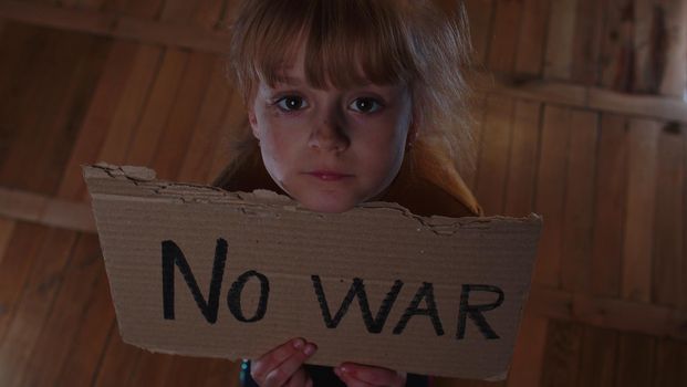 Afraid homeless toddler girl sitting holding inscription No War, hiding from bombing attack at home