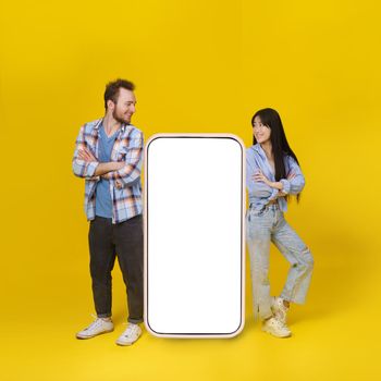 Asian girl and caucasian guy standing leaned on huge smartphone with white blank screen, mobile app advertisement and smile looking at each other isolated on yellow background. Product placement