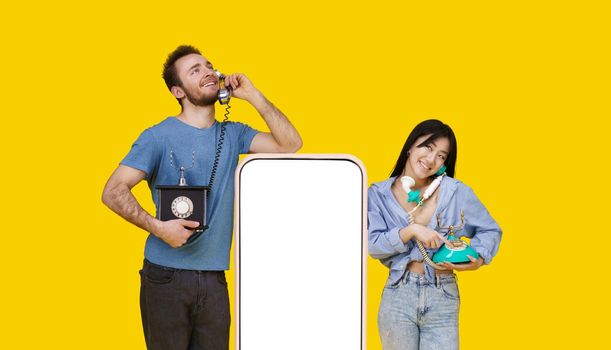 Asian girl and caucasian guy in love talking on vintage phones leaned on huge smartphone with blank screen, looking at each other isolated on yellow background. Product placement