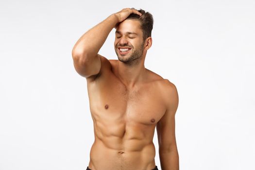 Wellbeing, health and bodybuilding concept. Handsome sexy young masculine guy with naked torso, touching hair and smiling with delightful expression, close eye, bragging good abs, six-pack body