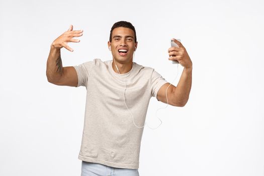 Sassy and arrogant cool hispanic man in t-shirt, shaking hands and dancing, singing along favourite songs, wear wired earphones, hold smartphone, enjoy nice music playlist, white background