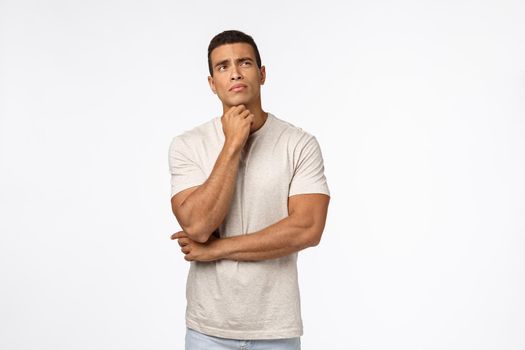 Indecisive young thoughtful male athlete trying make hard choice, decide what do, touching chin, looking up troubled, pondering, search answers in mind, standing white background thinking