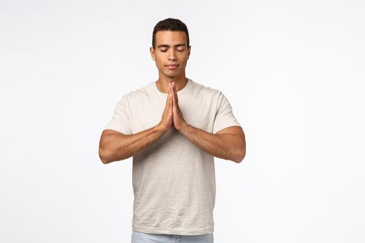 Handsome healthy muscular male in t-shirt, clasp hands together over chest, close eyes and meditate, practice yoga, praying with faithful happy expression, inhale fresh air, trying find peace in mind