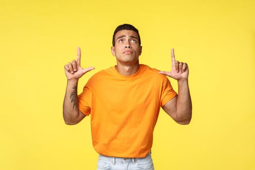 Young sad man with tattooed arm in orange t-shirt, white pants, looking up lonely with sadness and regret, pointing upwards, missed good sale, jealous of something, standing yellow background